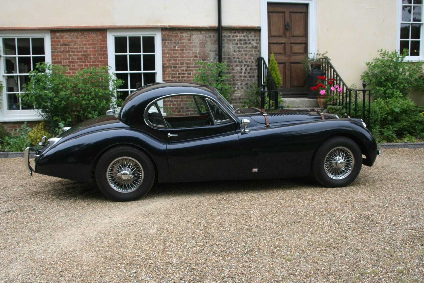 A black 1952 Jaguar XK120 fixed-head coupe with wire wheels parked on a gravel drive in front of a Georgian house