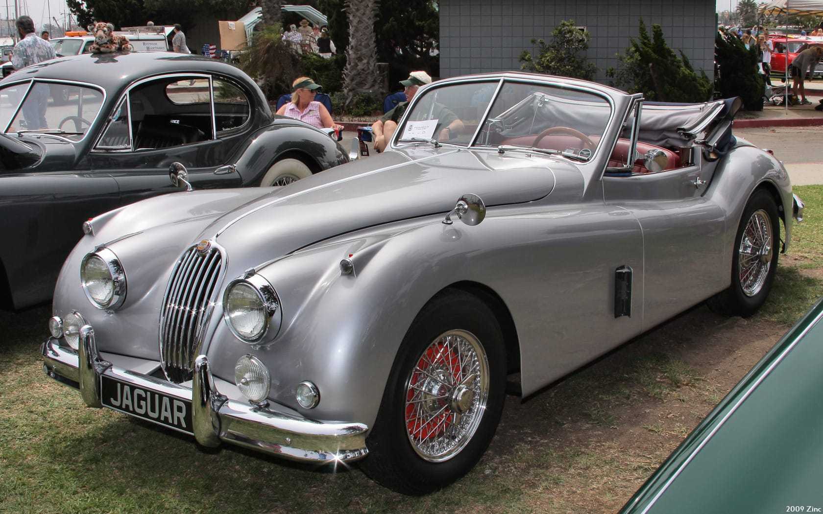 A silver 1956 Jaguar XK140 drophead coupe with its hood down on a showfield, front three-quarter view