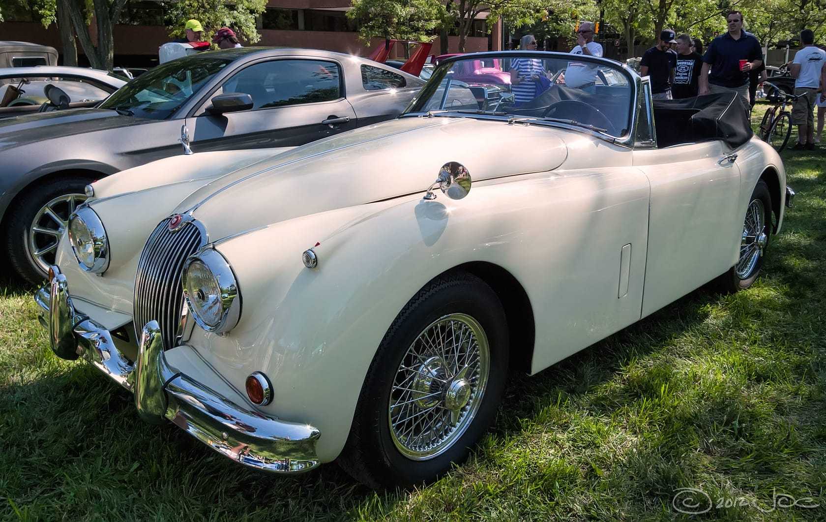 A cream 1958 Jaguar XK150 with its hood down on grass at a show, front three-quarter view
