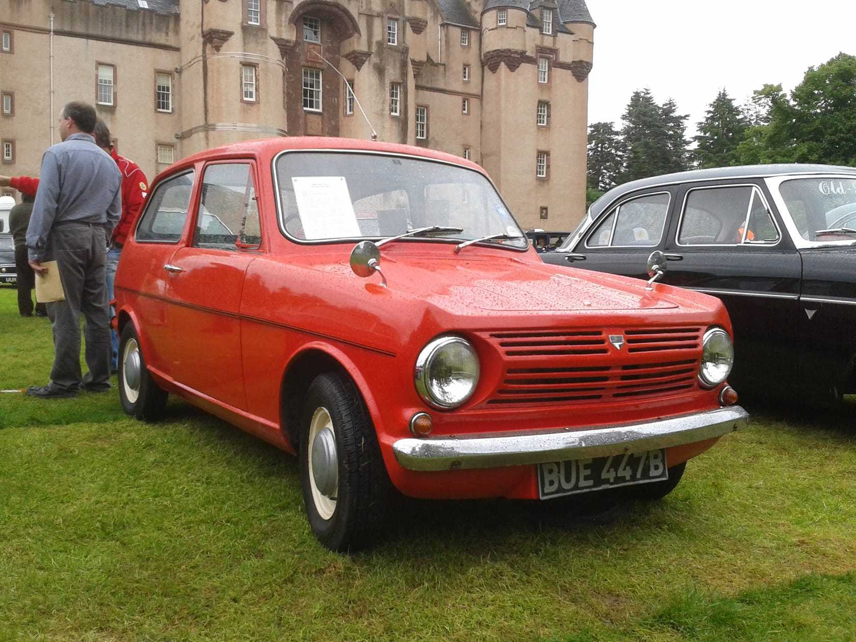 A red 1964 Reliant Rebel on grass at a show, front three-quarter view, UK registration BUE 447B