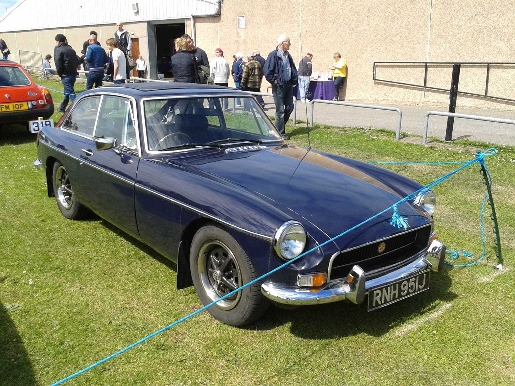 A red 1971 MGB GT with wire wheels parked on grass at a classic car gathering, viewed from the front three-quarter