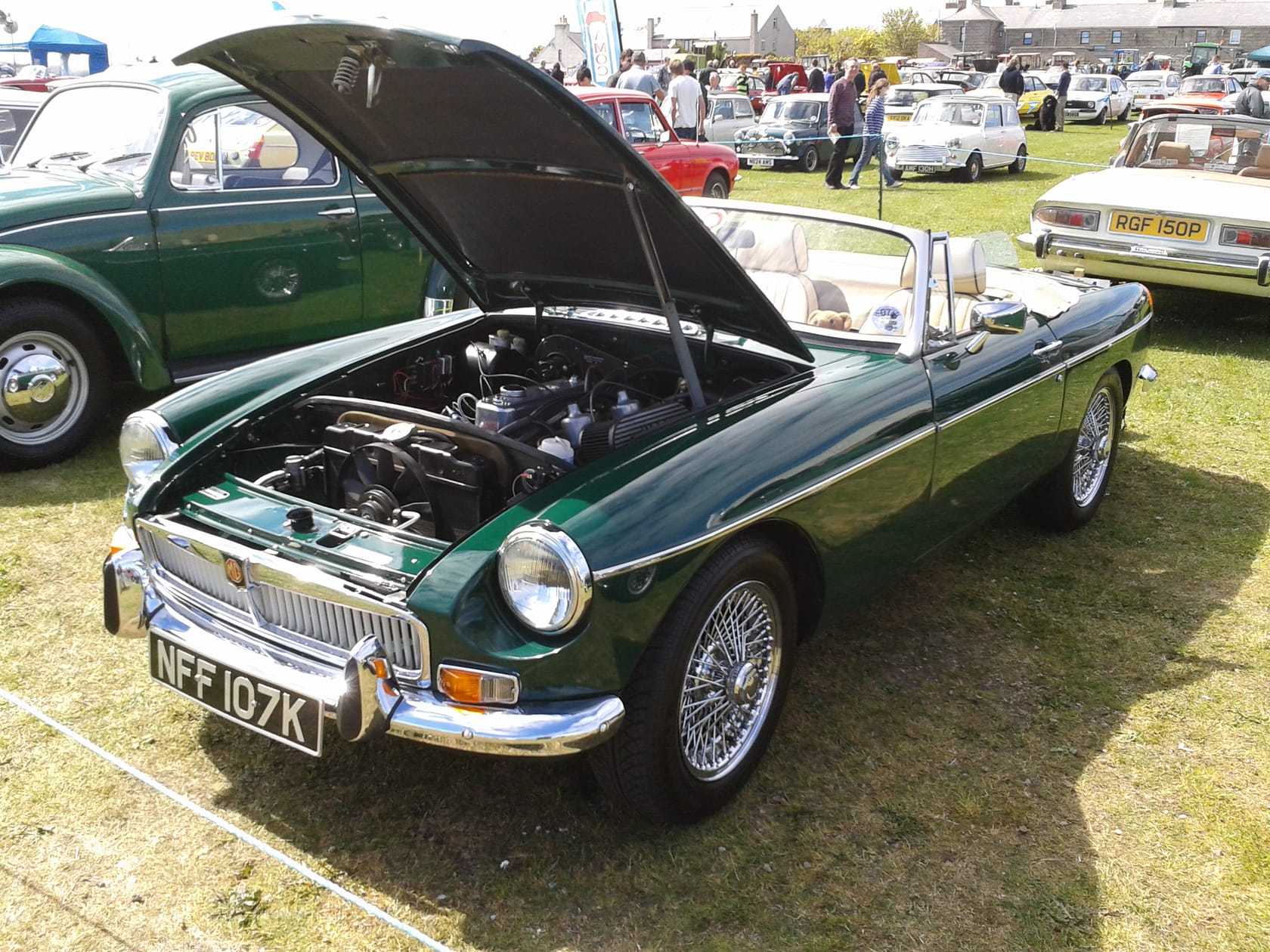 A British Racing Green MGB roadster with its bonnet open at a classic car show, showing the engine bay