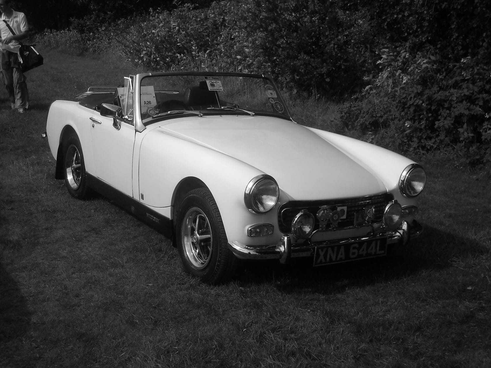 A bright blue 1973 MG Midget roadster with chrome bumpers and its hood down, parked on gravel outside a modern building
