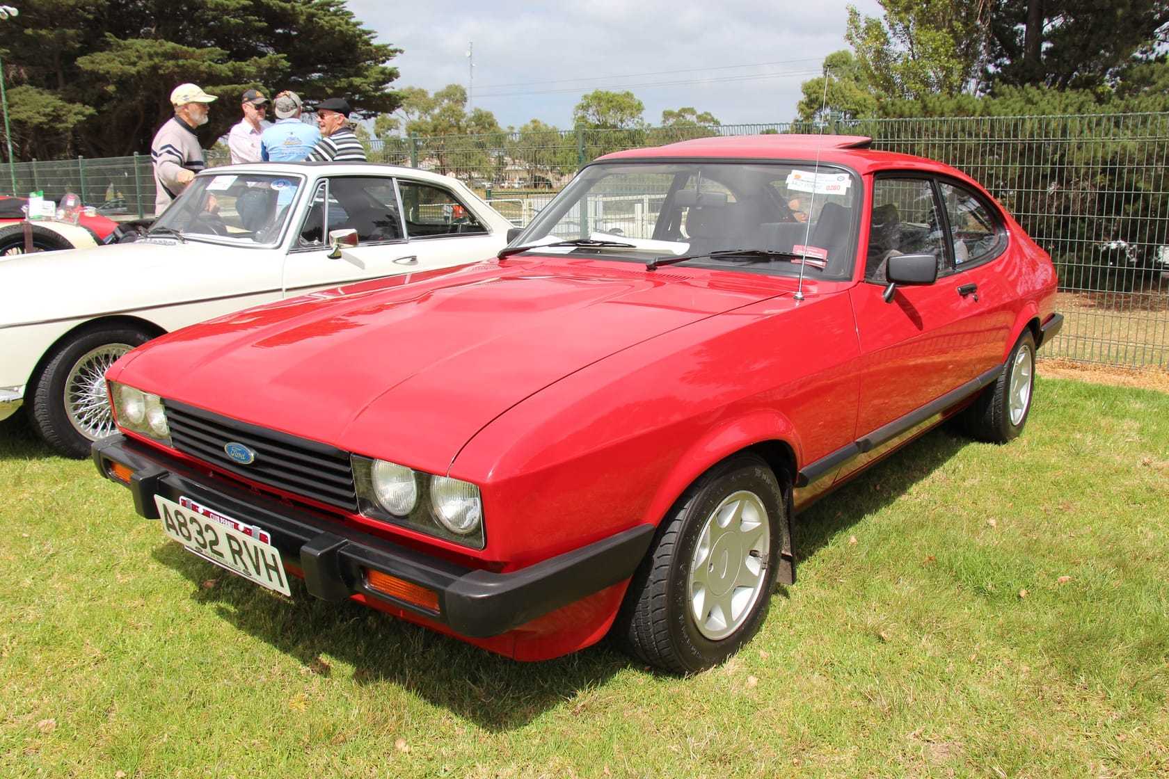 A red 1976 Ford Capri Mk3 with quad round headlamps parked on grass at a classic car show