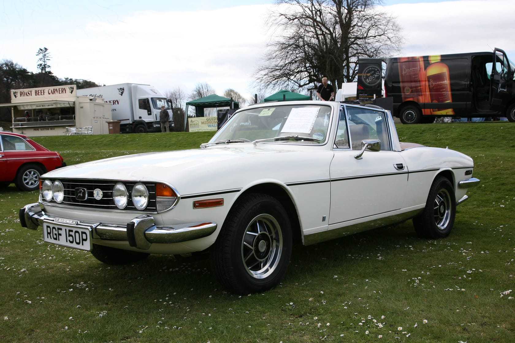 A white 1976 Triumph Stag convertible with its T-bar roll hoop and hood down, parked on grass at a show