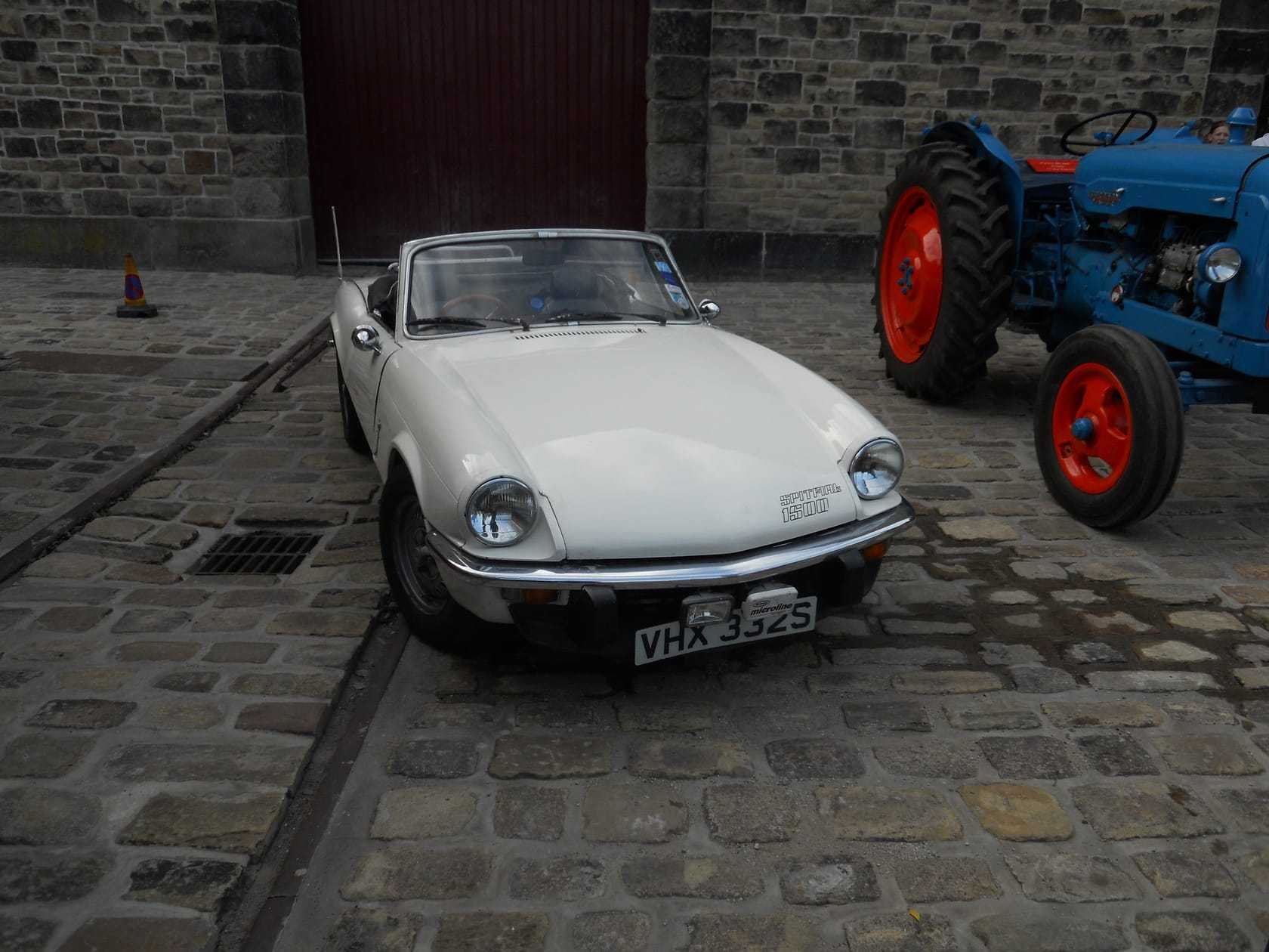 A dark green 1978 Triumph Spitfire 1500 roadster with its hood down parked on tarmac in front of a park noticeboard