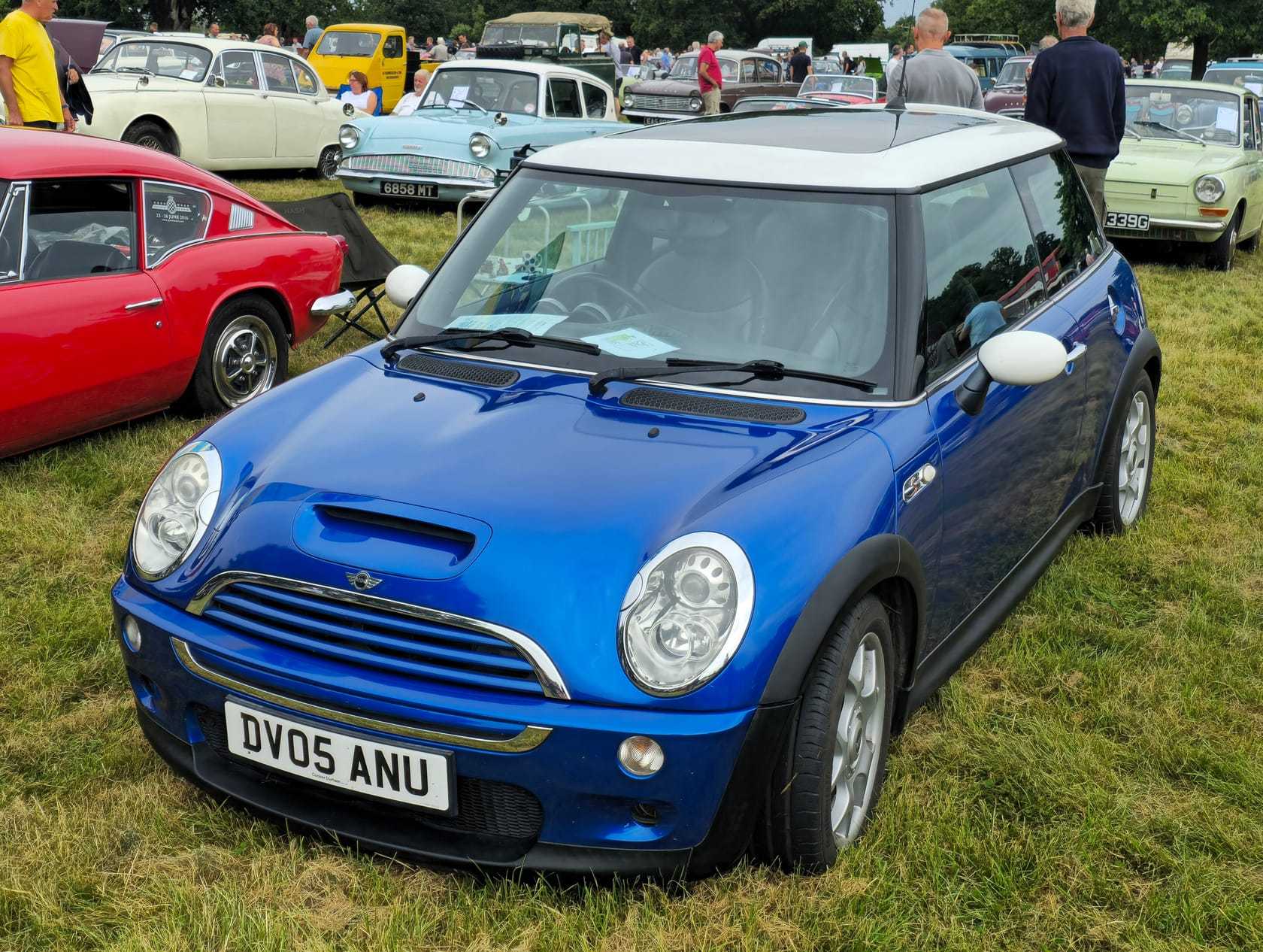 A blue 2005 MINI Cooper S R53 with a white roof and a UK registration on a classic car show field, classic cars behind
