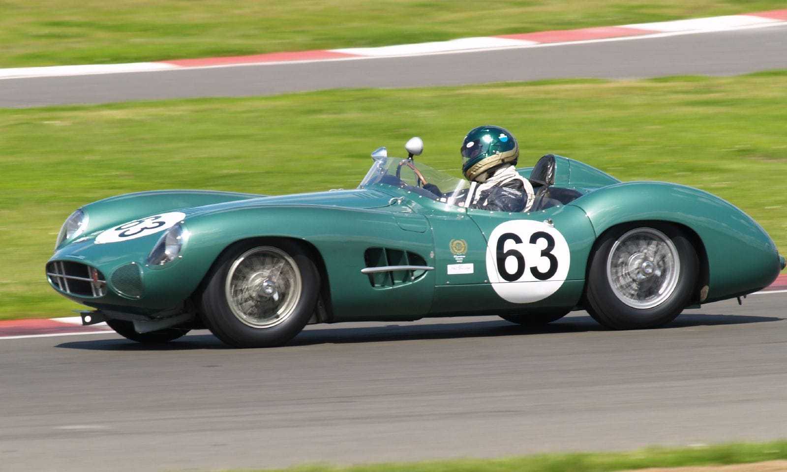 A green Aston Martin DBR1 sports racing car with the number 63, driven at speed on a circuit by a driver in a green helmet
