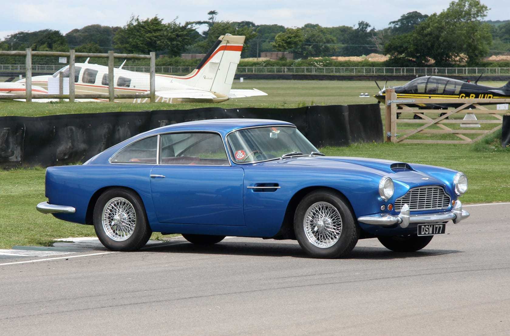 A blue Aston Martin DB4 on a race circuit at Goodwood with a light aircraft behind