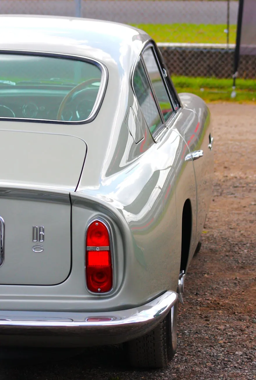 The rear three-quarter of a silver Aston Martin DB6, its "DB6" bootlid badge visible, parked on gravel by a circuit fence