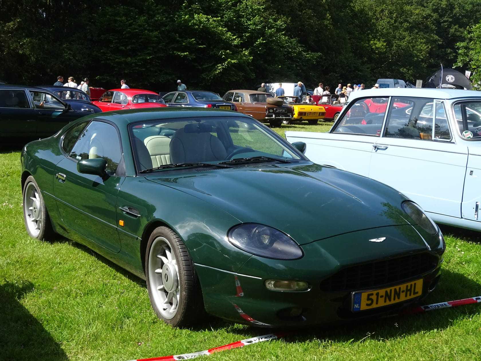 A dark green Aston Martin DB7 parked on grass among other classics at a club gathering with people browsing behind