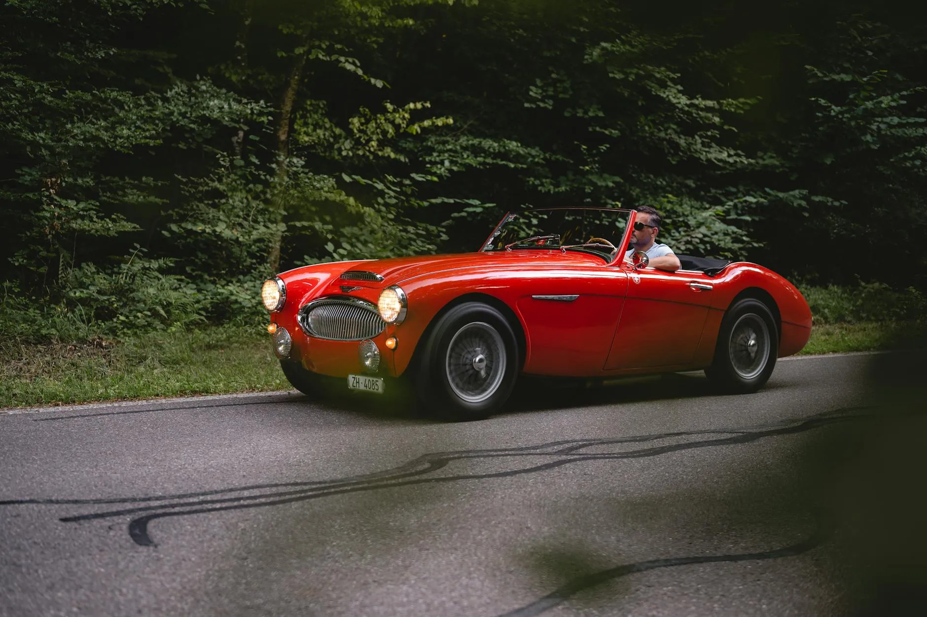 A red Austin-Healey 3000 roadster being driven on a forest road with its headlamps lit