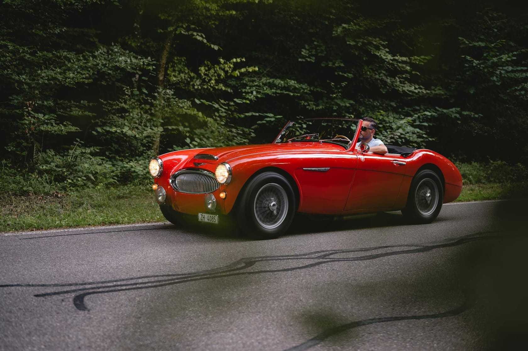 A red Austin-Healey 3000 roadster being driven on a forest road with its headlamps lit