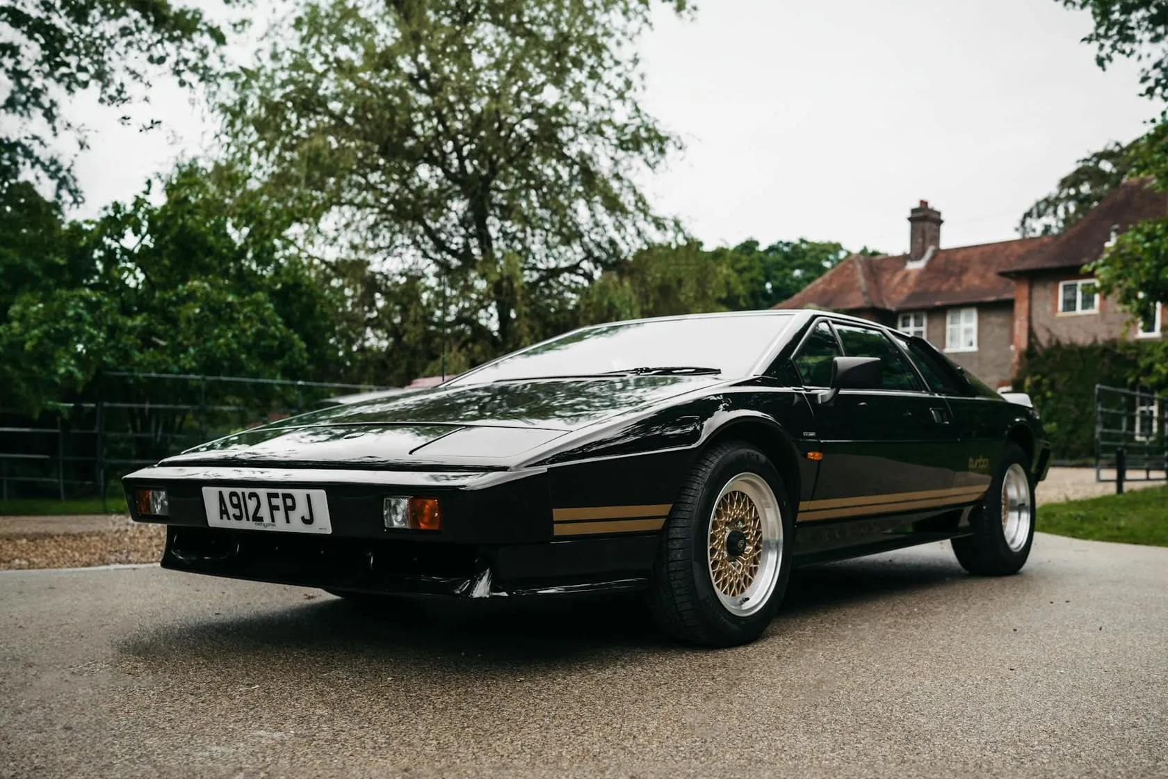 A black Lotus Esprit with a gold side stripe and an A-suffix number plate, parked on a driveway in front of a country house