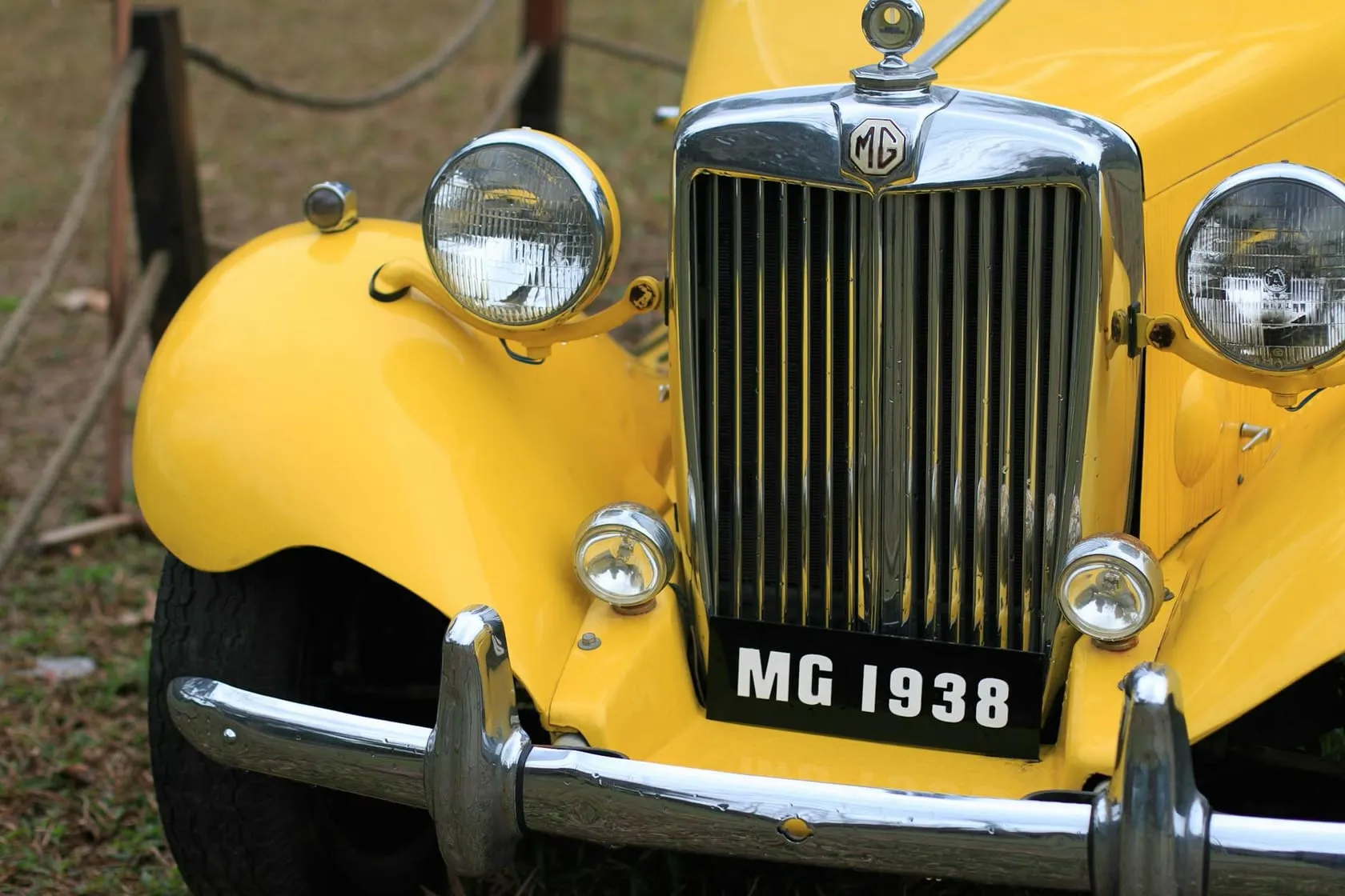 The chrome grille and twin headlamps of a yellow MG T-type, with an "MG 1938" number plate