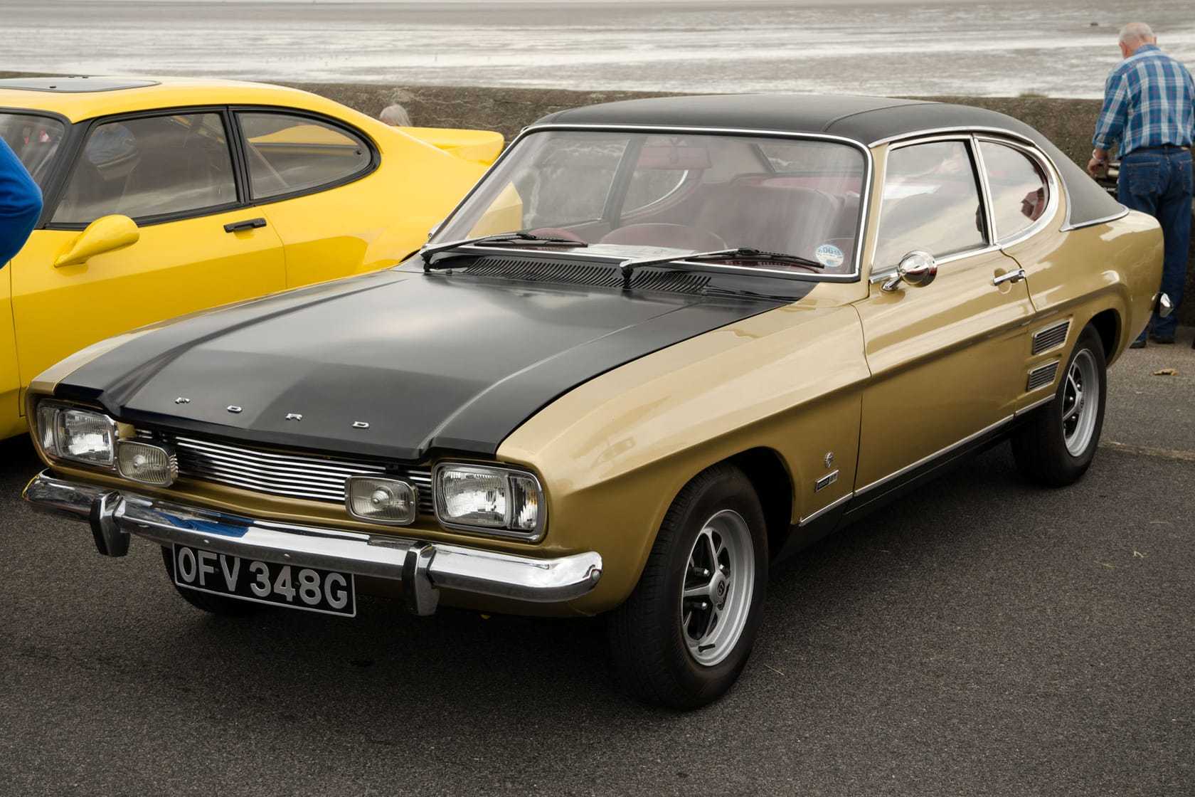 A gold 1969 Ford Capri Mk1 2000GT with a black bonnet and a UK registration parked by the sea