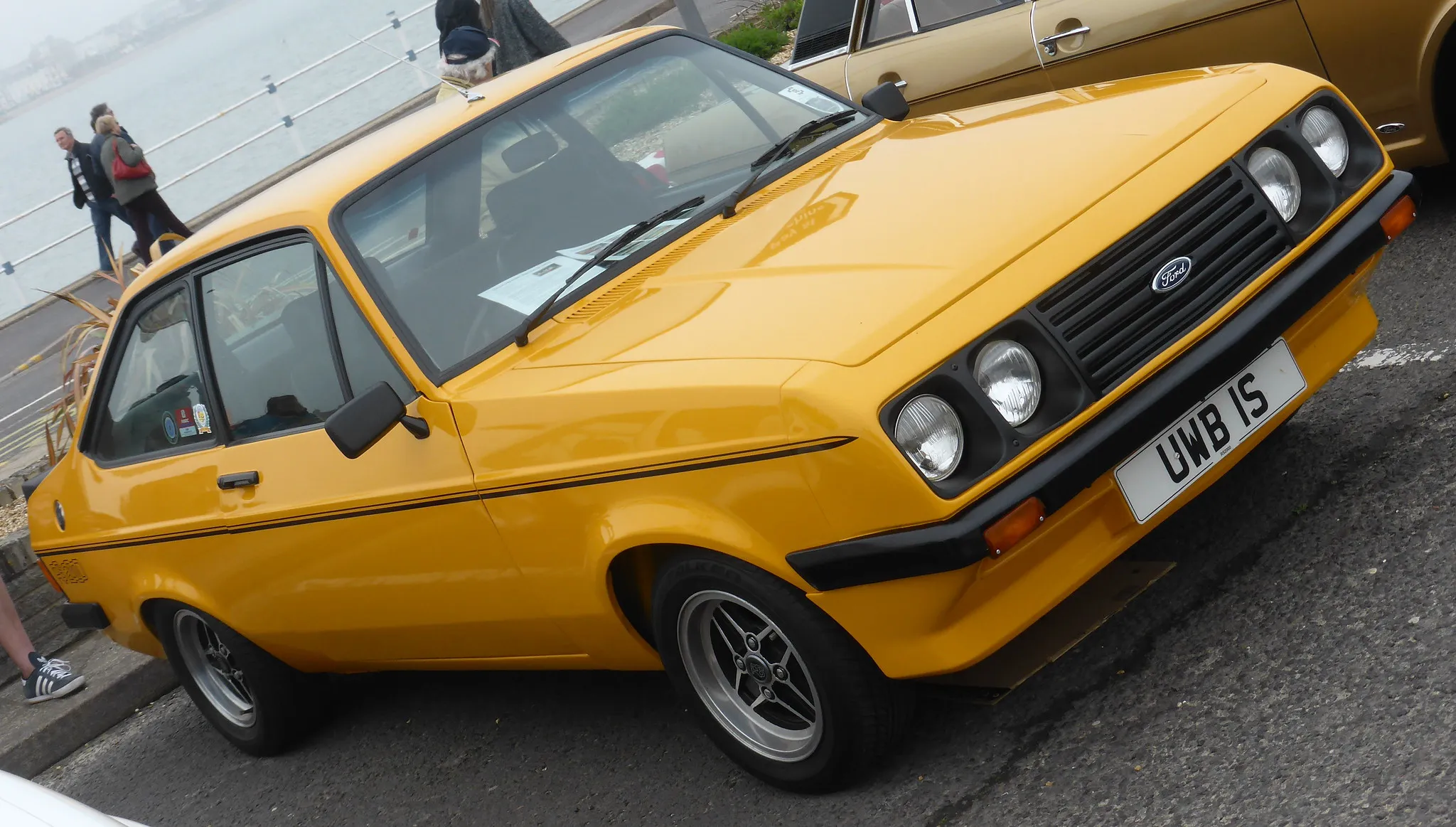 A yellow Ford Escort Mk2 RS2000 with its black polyurethane droop-snoot nose, front three-quarter view at a seafront classic car gathering