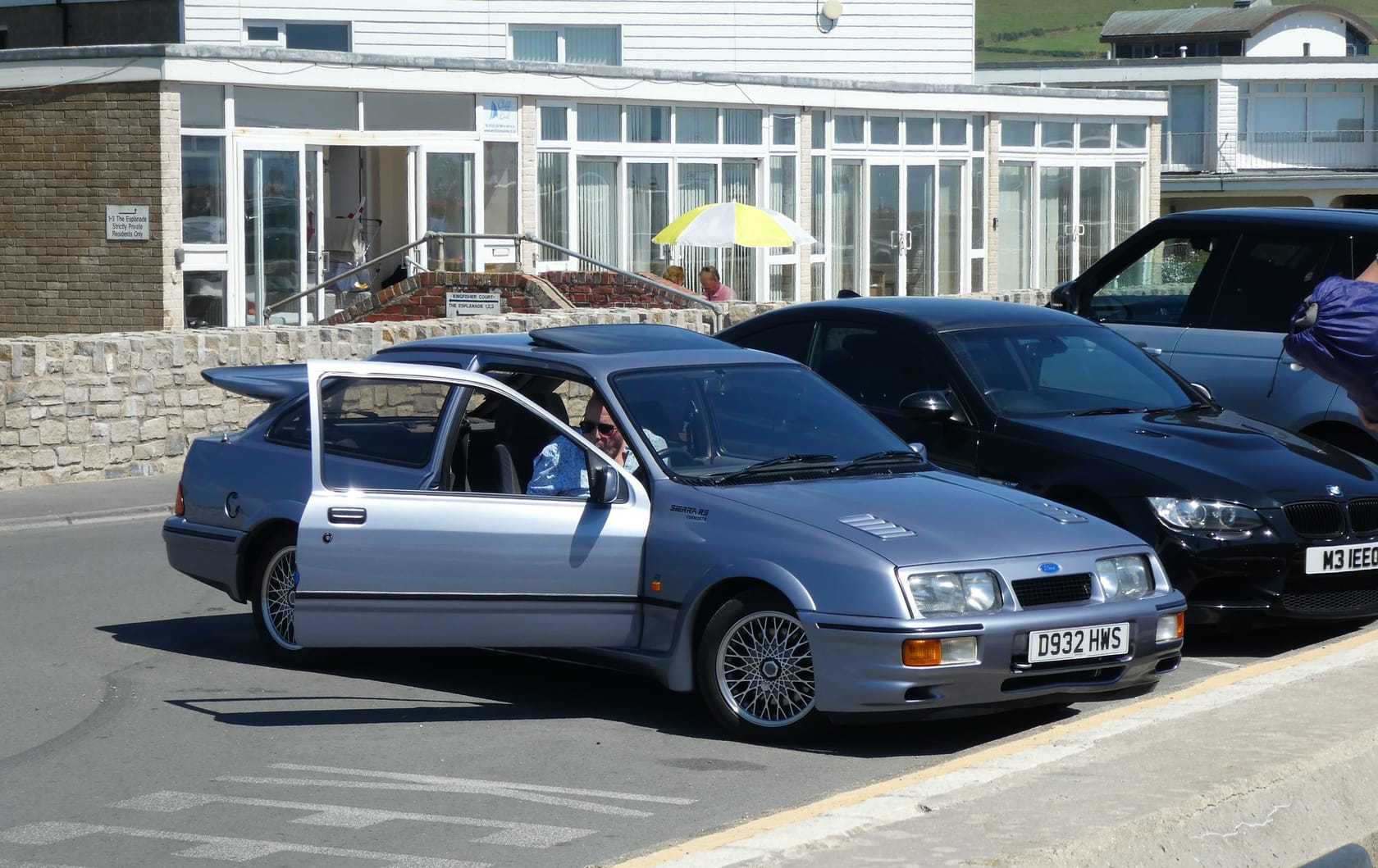 A silver Ford Sierra RS Cosworth three-door with its whale-tail spoiler and a UK registration, parked on a British seafront