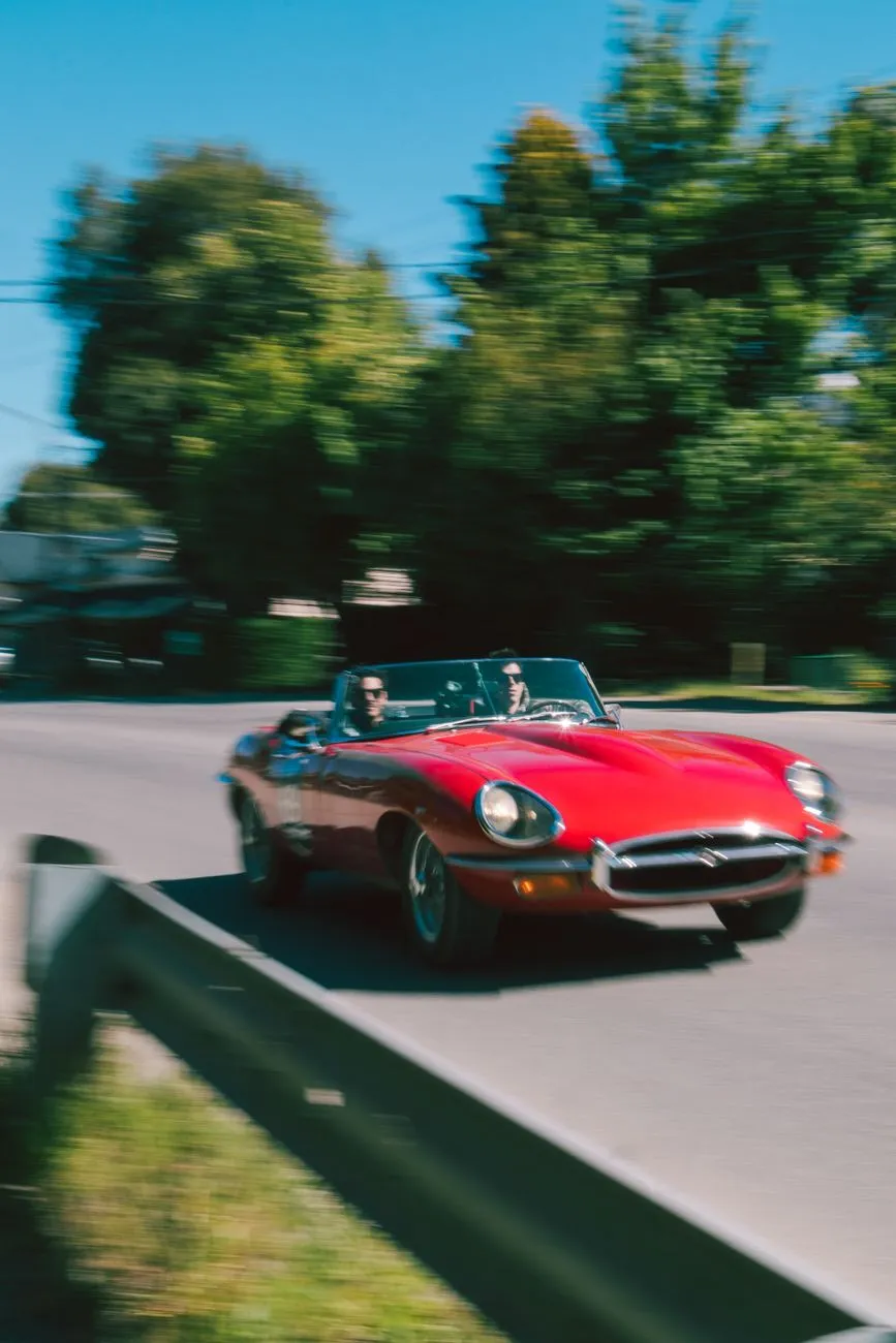 A red Jaguar E-Type roadster being driven along a sunny tree-lined road, front three-quarter view