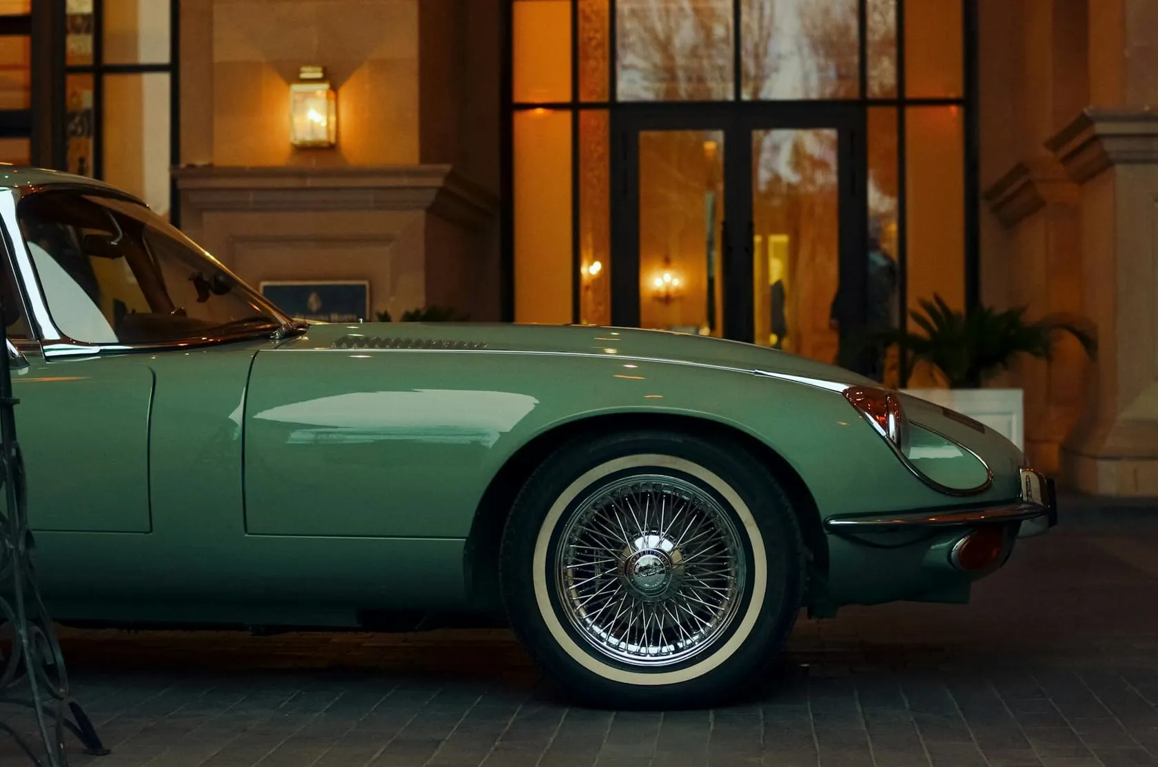 A sage-green Jaguar E-Type coupe in side profile, with chrome wire wheels and whitewall tyres, parked outside a lamplit hotel entrance at dusk