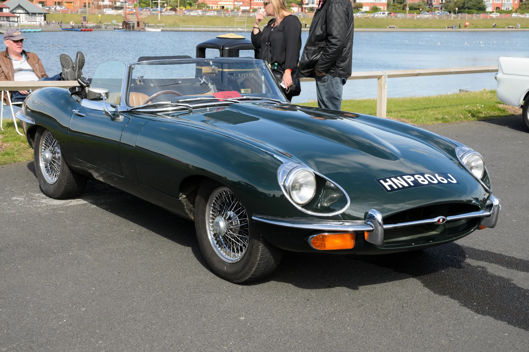 A dark green Jaguar E-Type Series 2 roadster with open headlamps parked beside water