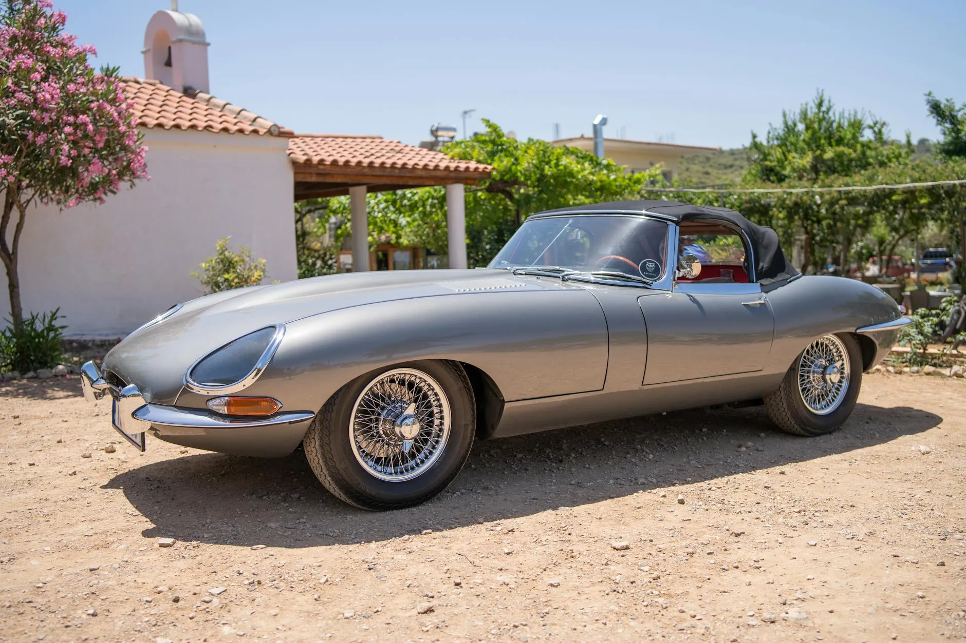 A silver Jaguar E-Type Series 1 roadster with covered headlamps and wire wheels parked in a sunny yard, side view