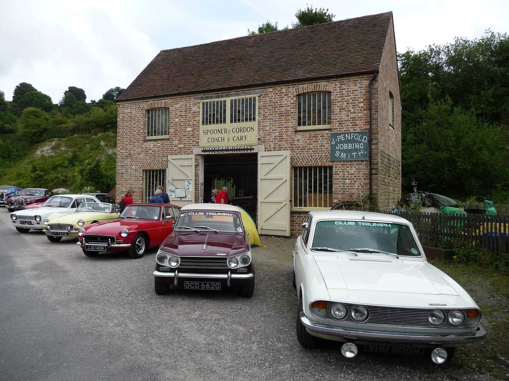 A lineup of classic British cars at a show, a Jaguar, a Ford, a red MGB and two Triumphs, parked in front of an old brick workshop, all on UK plates