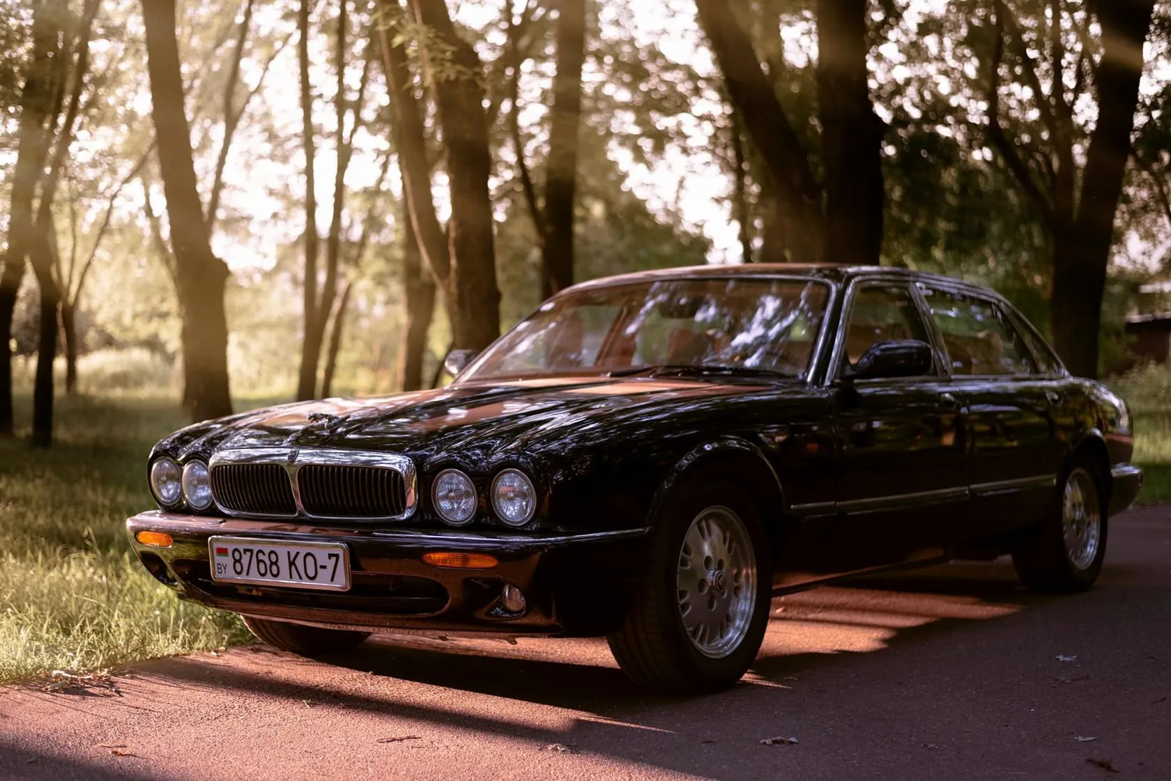 A black 1990s Jaguar XJ saloon, the four-headlamp X300 shape, parked on a tree-lined road in golden evening light