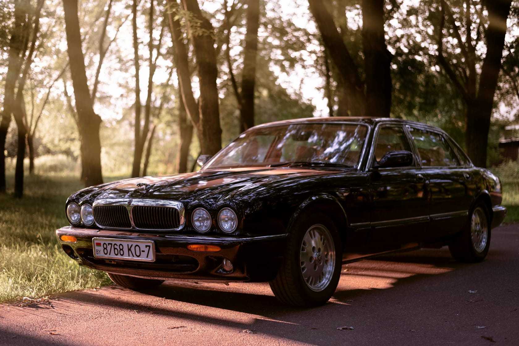 A black 1990s Jaguar XJ saloon, the four-headlamp X300 shape, parked on a tree-lined road in golden evening light