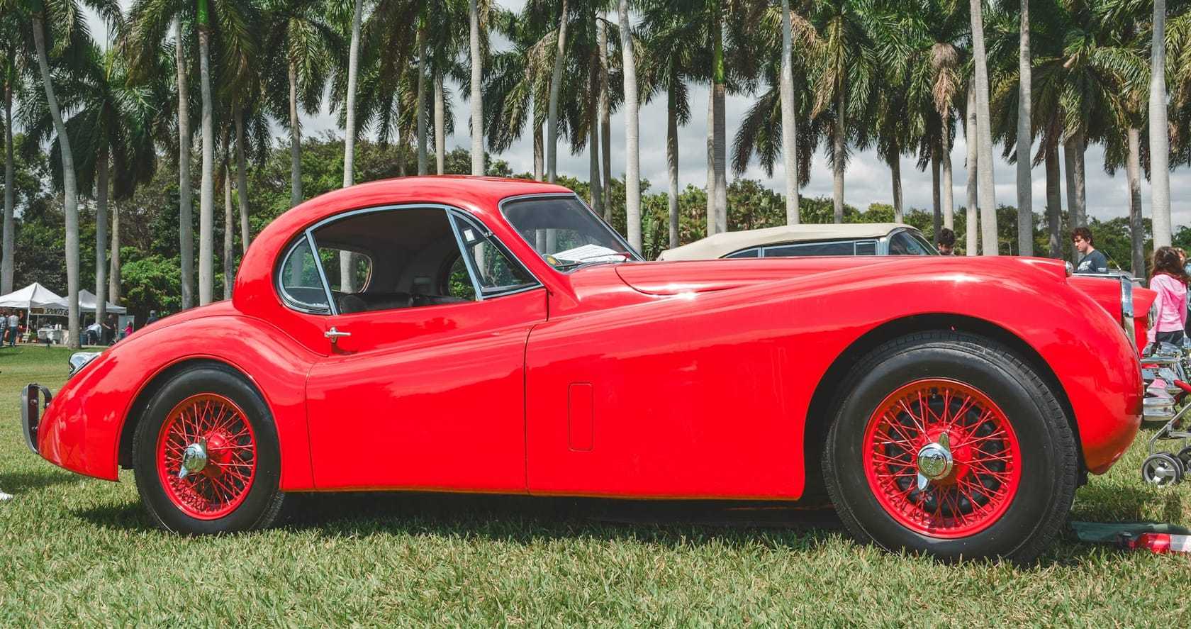 A red Jaguar XK120 fixed-head coupe in side profile with red wire wheels, parked on grass with palm trees behind