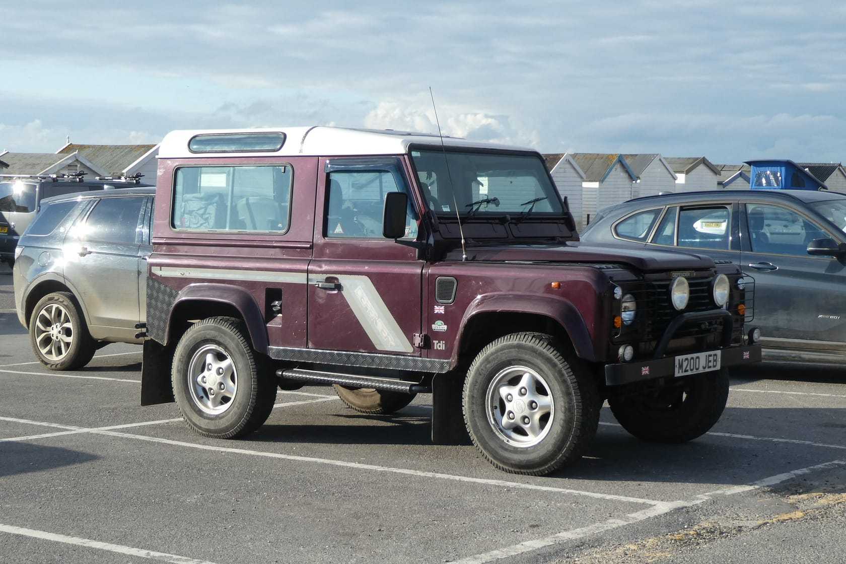 A dark red Land Rover Defender 90 County with a white roof parked at the coast, wearing a UK number plate