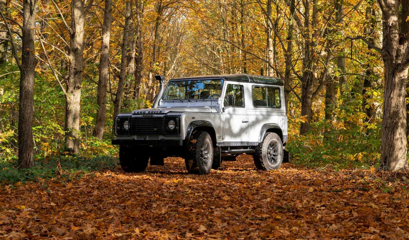 A silver Land Rover Defender 110 parked on a leaf-strewn track in an autumn forest