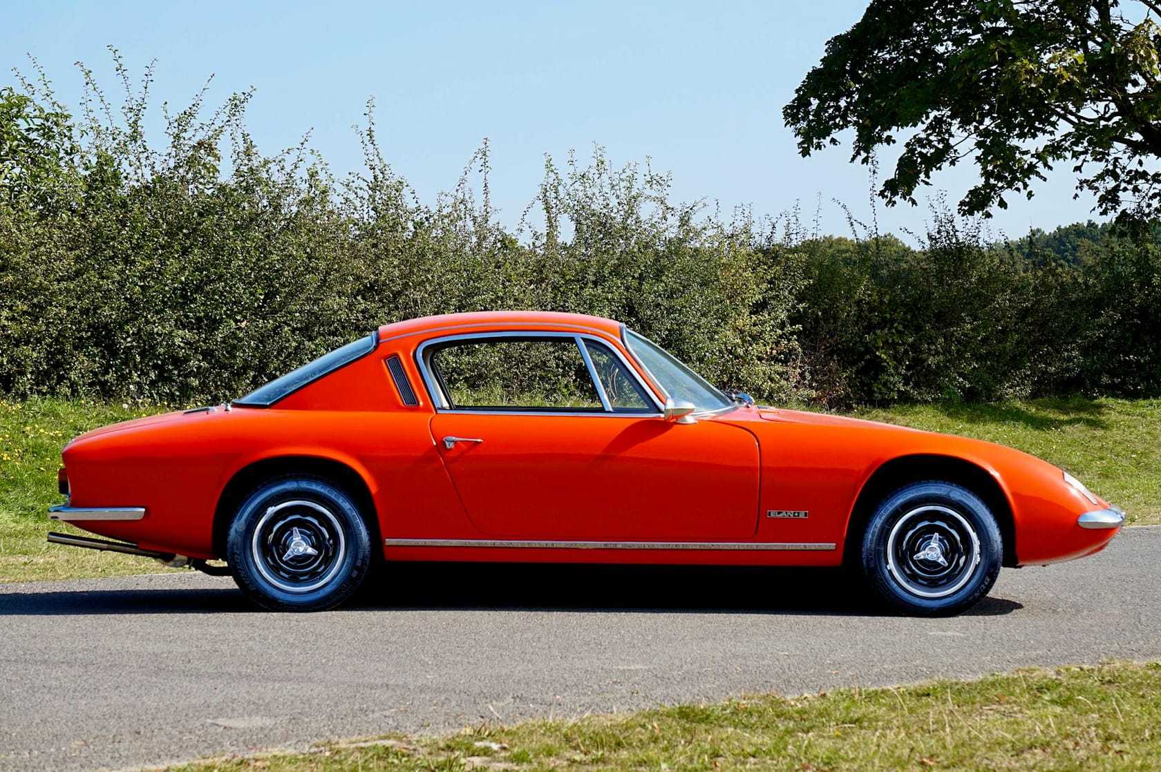 An orange Lotus Elan +2 fixed-head coupe in side profile against a hedge