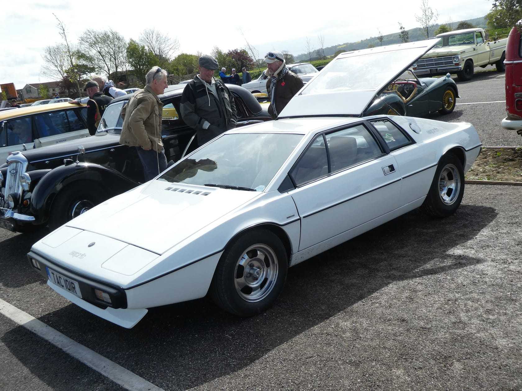 A white 1977 Lotus Esprit S1 with a UK registration on a show field, a pre-war car beside it