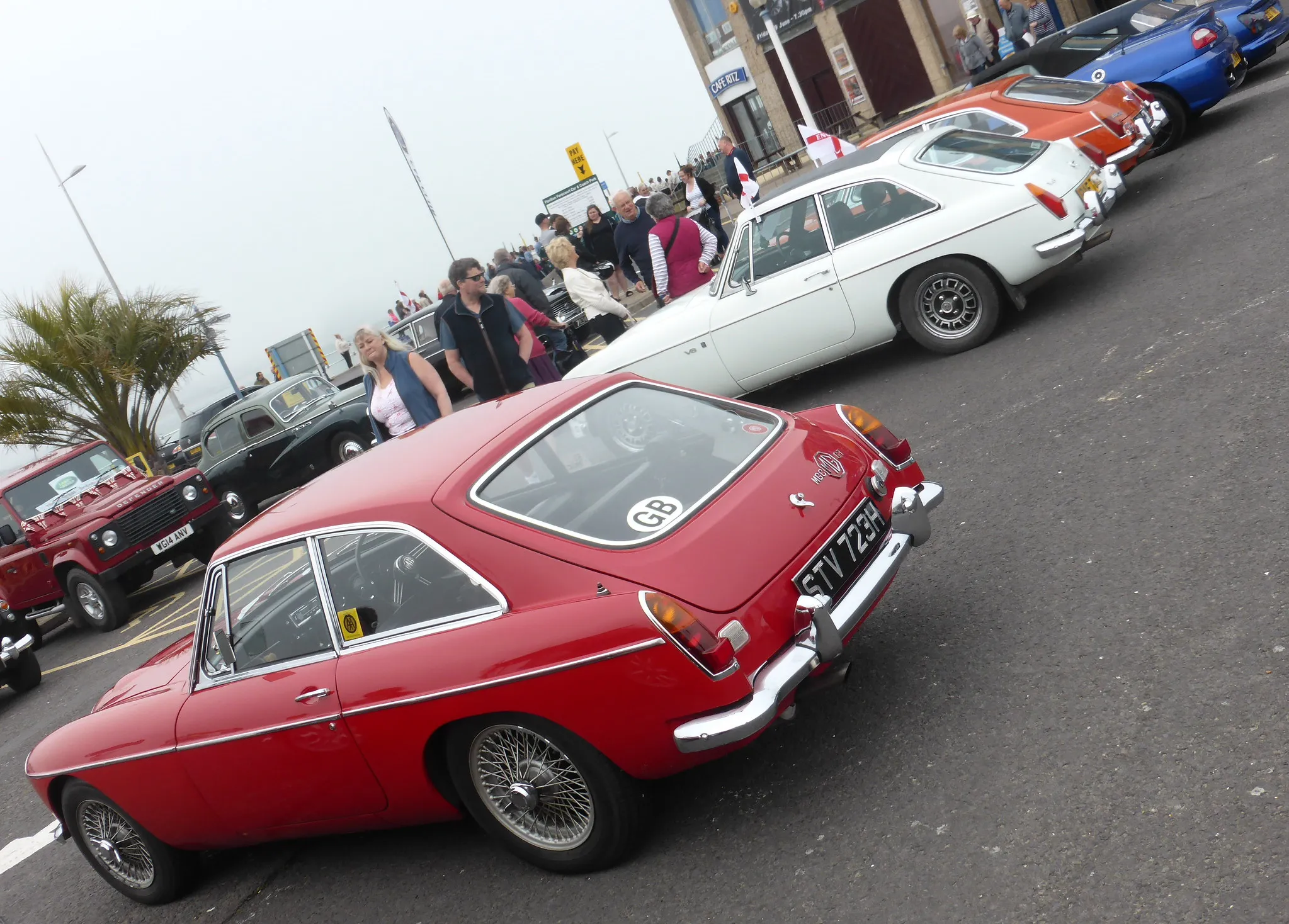 A row of classic MG sports cars, led by a red MGB GT, at a seaside MG gathering
