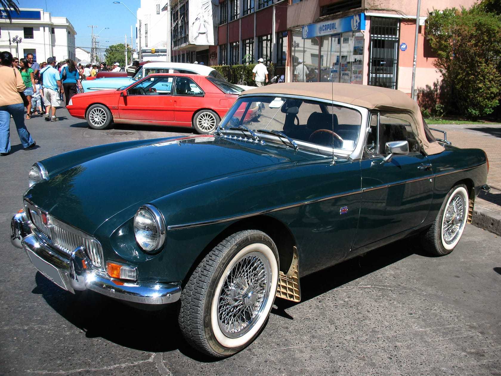 A dark green MGB roadster with wire wheels and whitewall tyres parked at a street gathering overseas