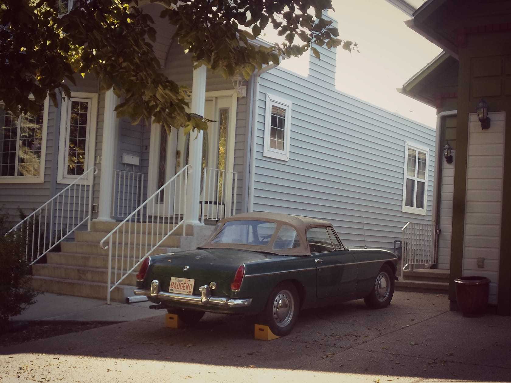 A dark MGB roadster with its hood up parked on the drive of a clapboard house overseas