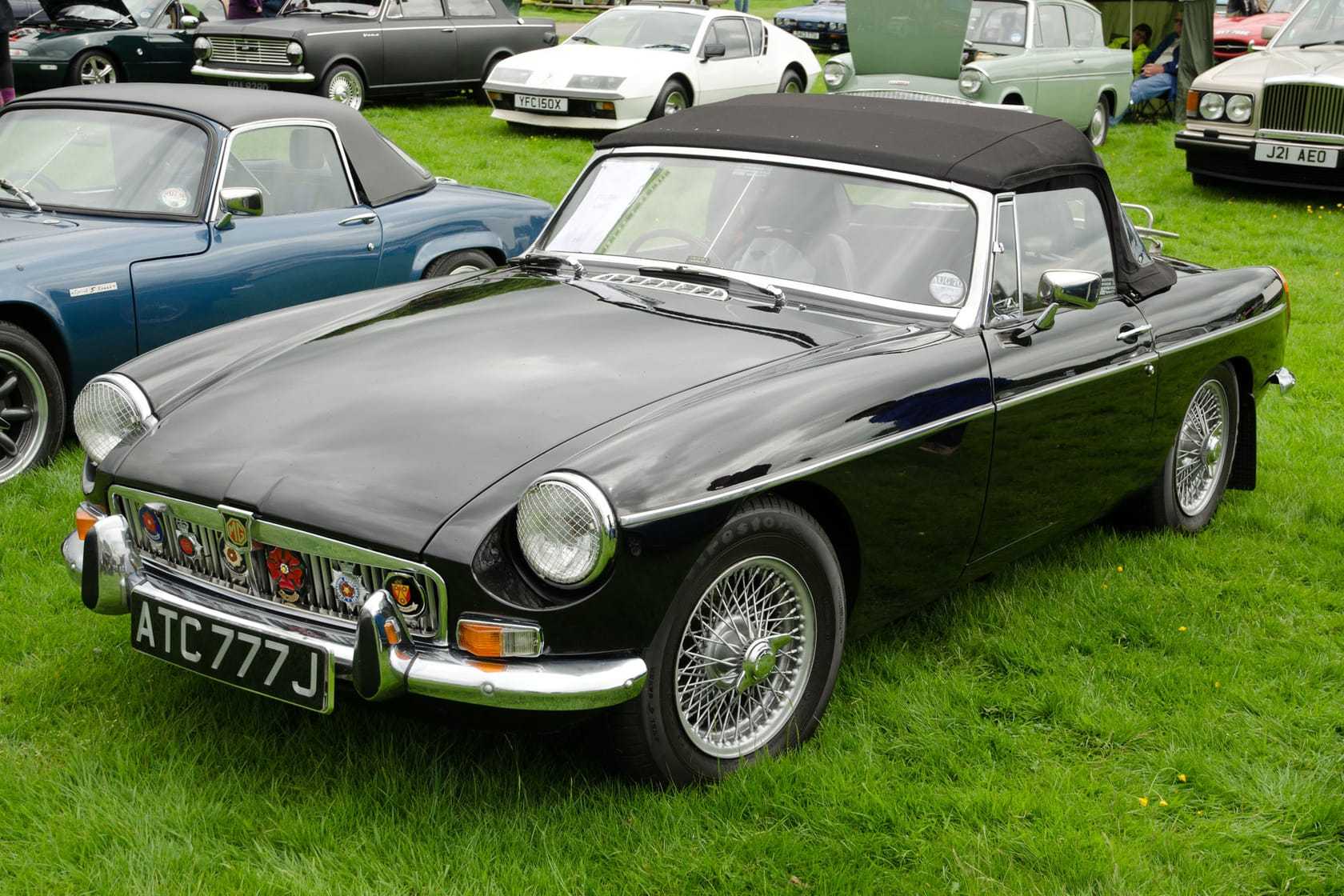 A black 1970 chrome-bumper MG MGB roadster with wire wheels and its hood up, parked on a show field