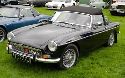 A black 1970 chrome-bumper MG MGB roadster with wire wheels and its hood up, parked on a show field