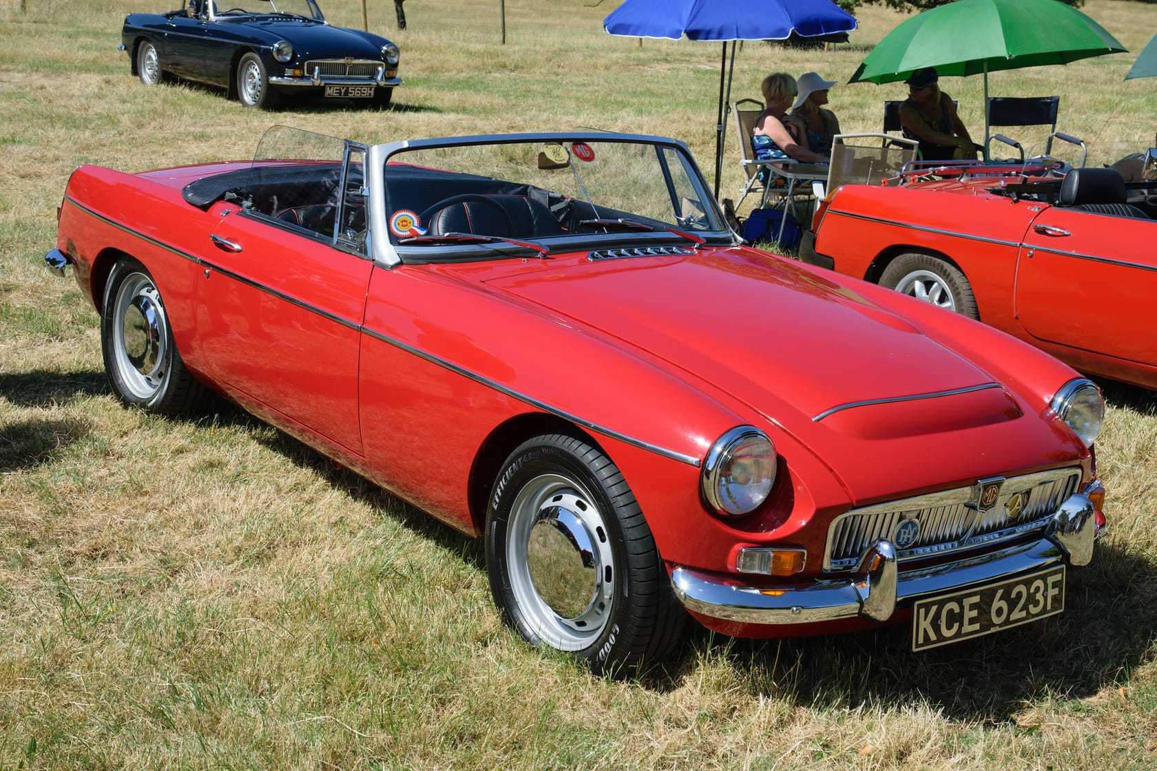 A red 1968 MGC roadster with its hood down and a UK registration on a show field