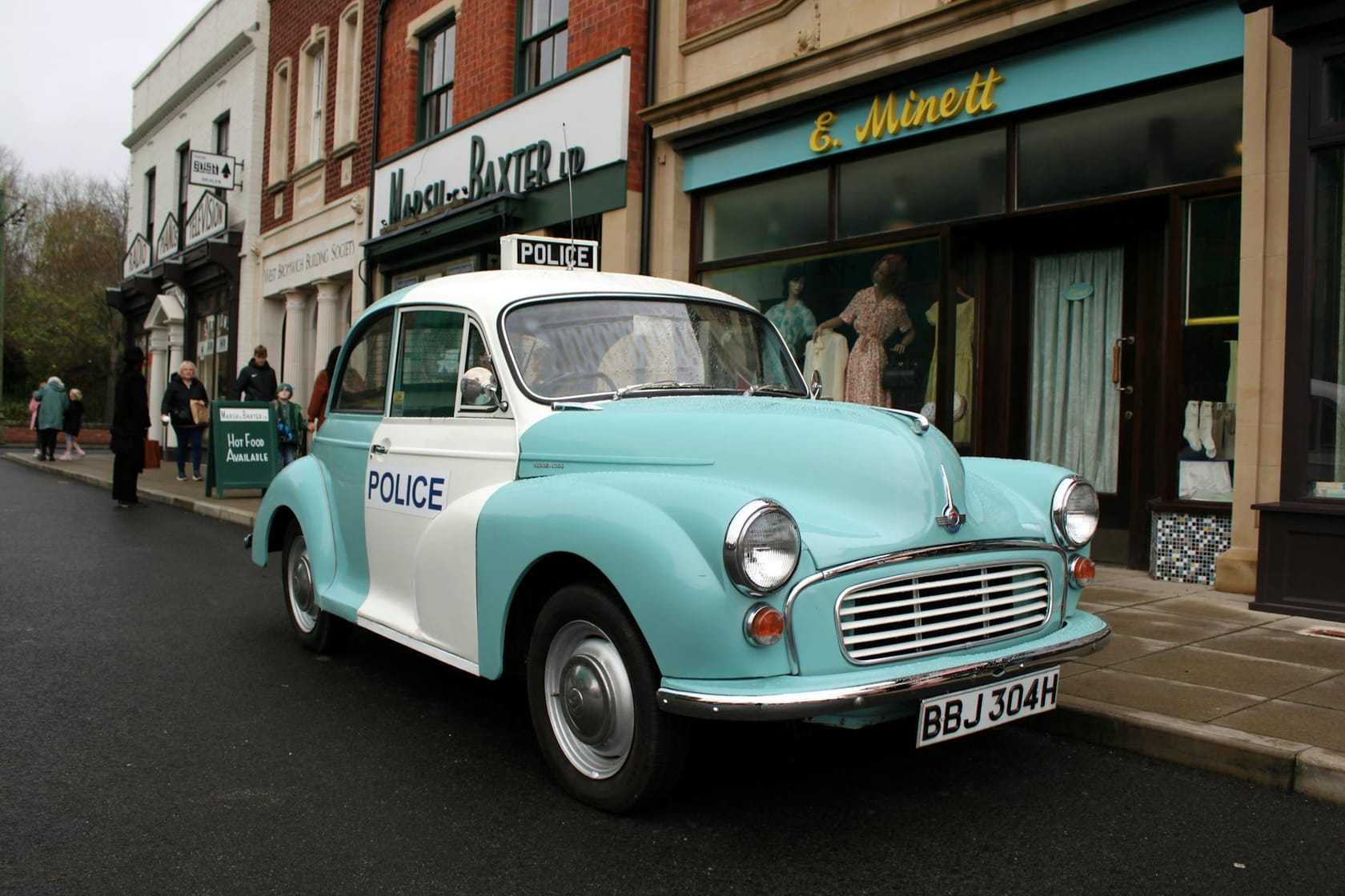 A pale blue and white Morris Minor police panda car parked on a British high street