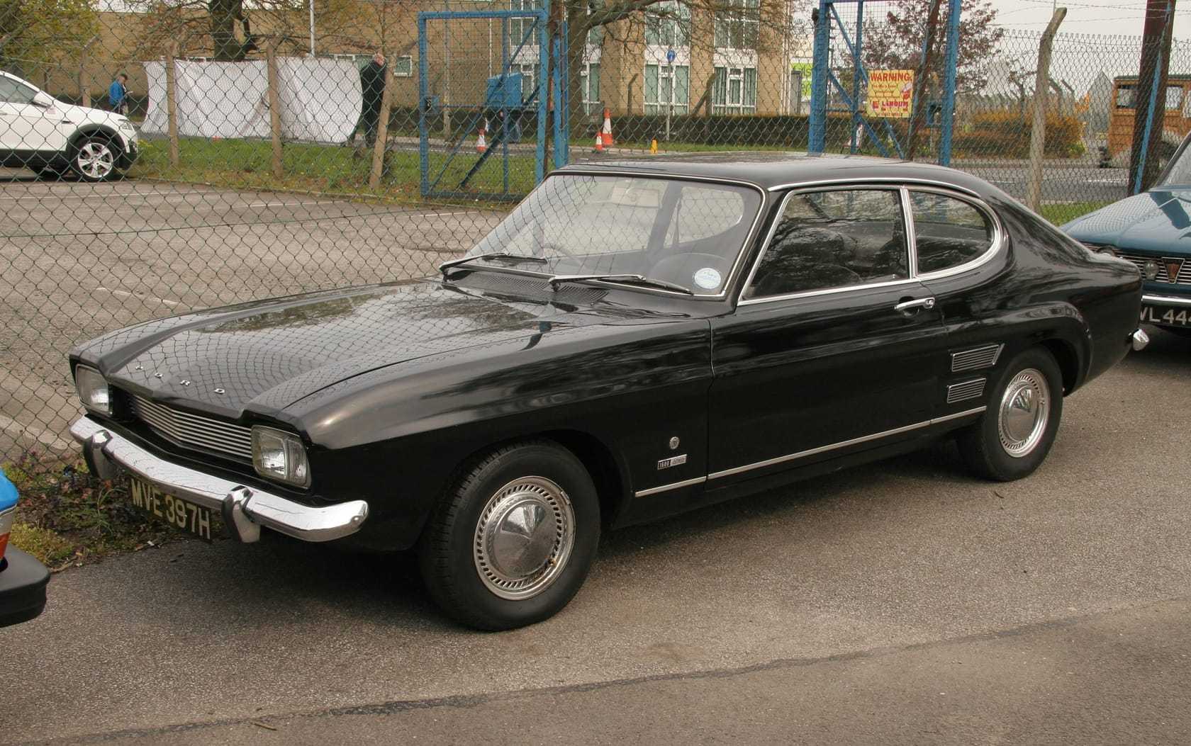 A black 1969 Ford Capri Mk1 1600XL with chrome hubcaps parked on a tarmac yard beside a chain-link fence, UK registration MVE 397H