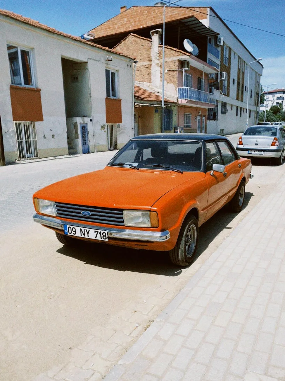 An orange Ford Cortina Mk4 four-door saloon parked on a sunlit street, front three-quarter view