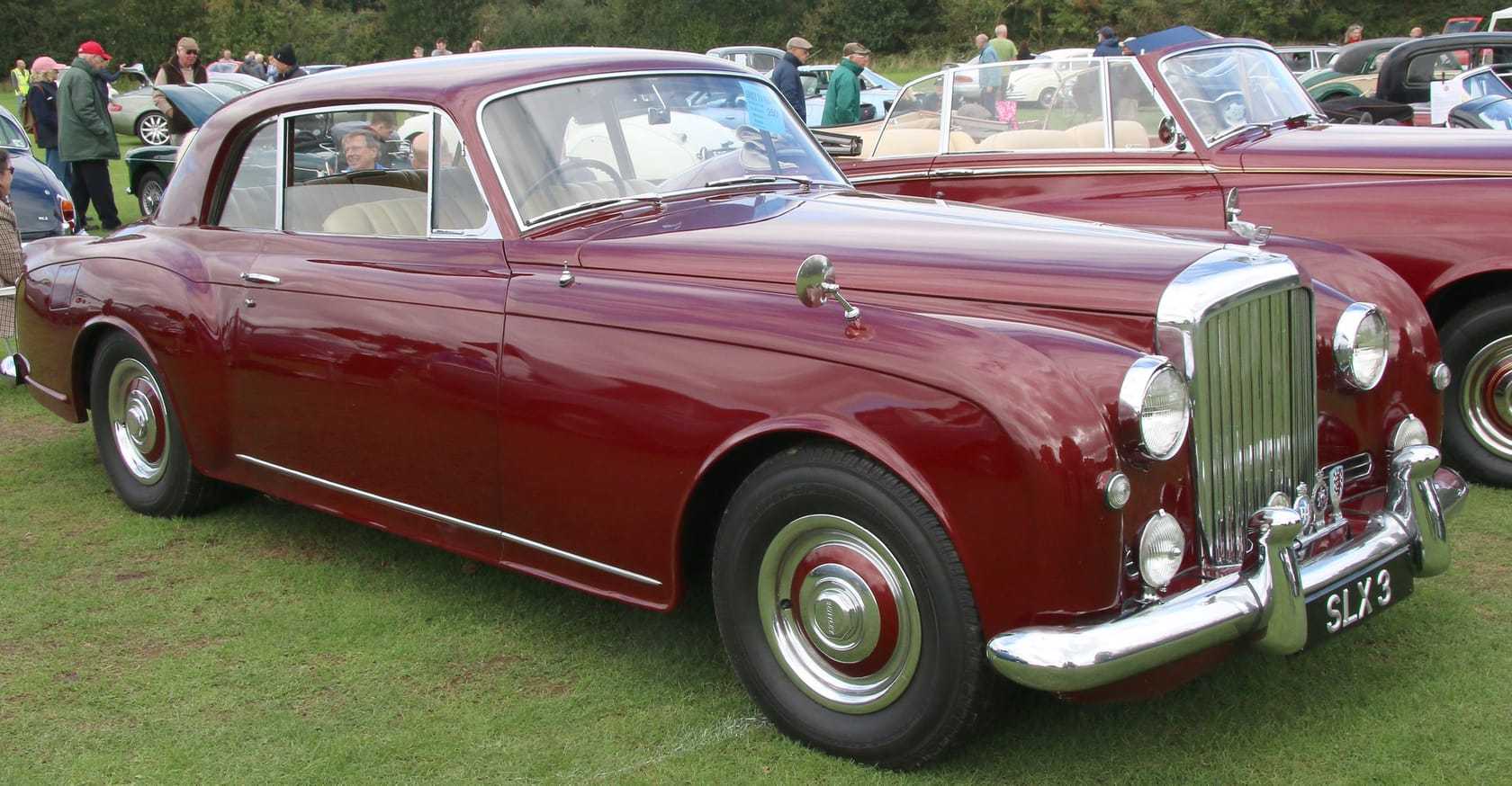 A maroon 1956 Bentley S1 Continental coupe by Park Ward parked on grass at a British show, three-quarter front view
