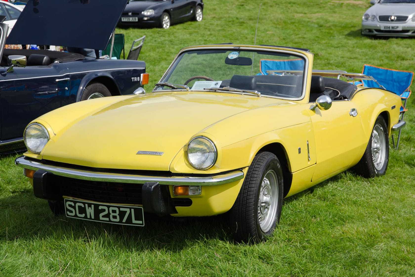 A yellow 1973 Triumph Spitfire Mk4 with a UK registration on a show field, its hood down