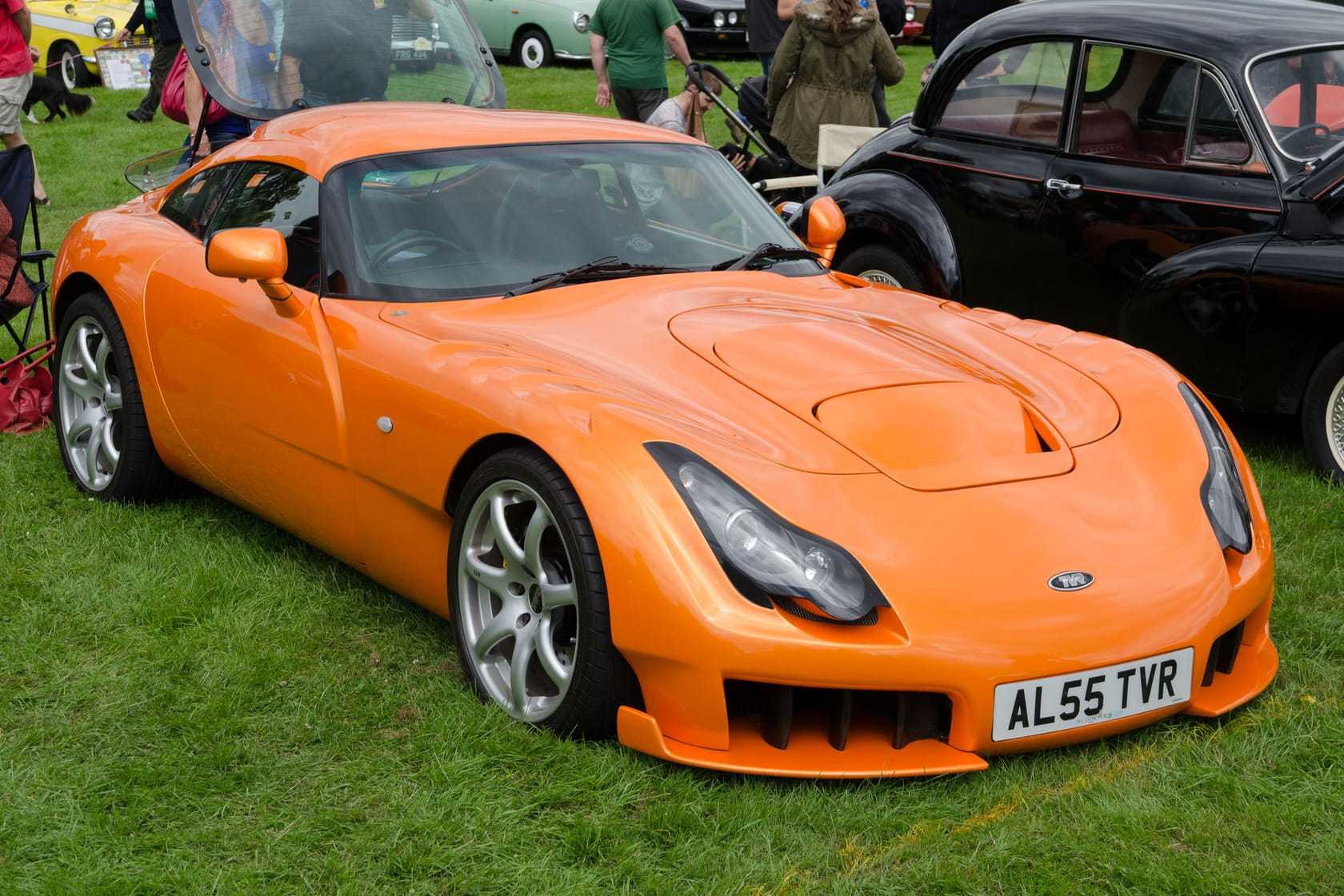An orange 2005 TVR Sagaris with a UK registration parked on a show field beside a black pre-war saloon