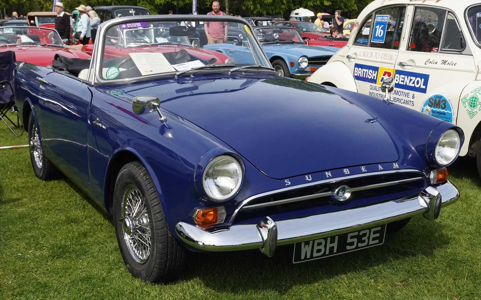 A dark blue Sunbeam Alpine Series V with wire wheels on grass at a show, front three-quarter view, UK registration WBH 53E
