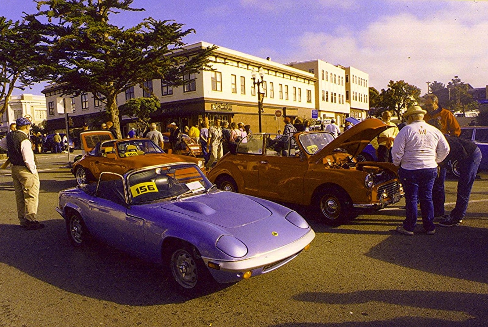 A stylised period illustration of a purple Lotus Elan roadster parked on a 1960s street with onlookers