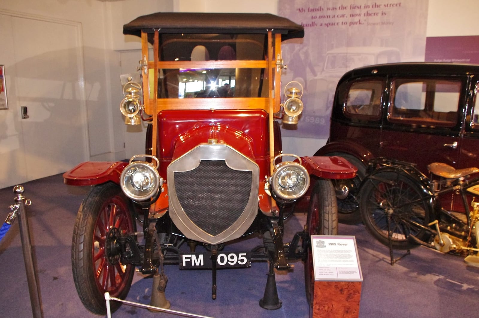 A red 1909 Rover 15 with brass lamps and a wooden-framed cab on museum display beside an early Rover motorcycle, UK registration FM 095