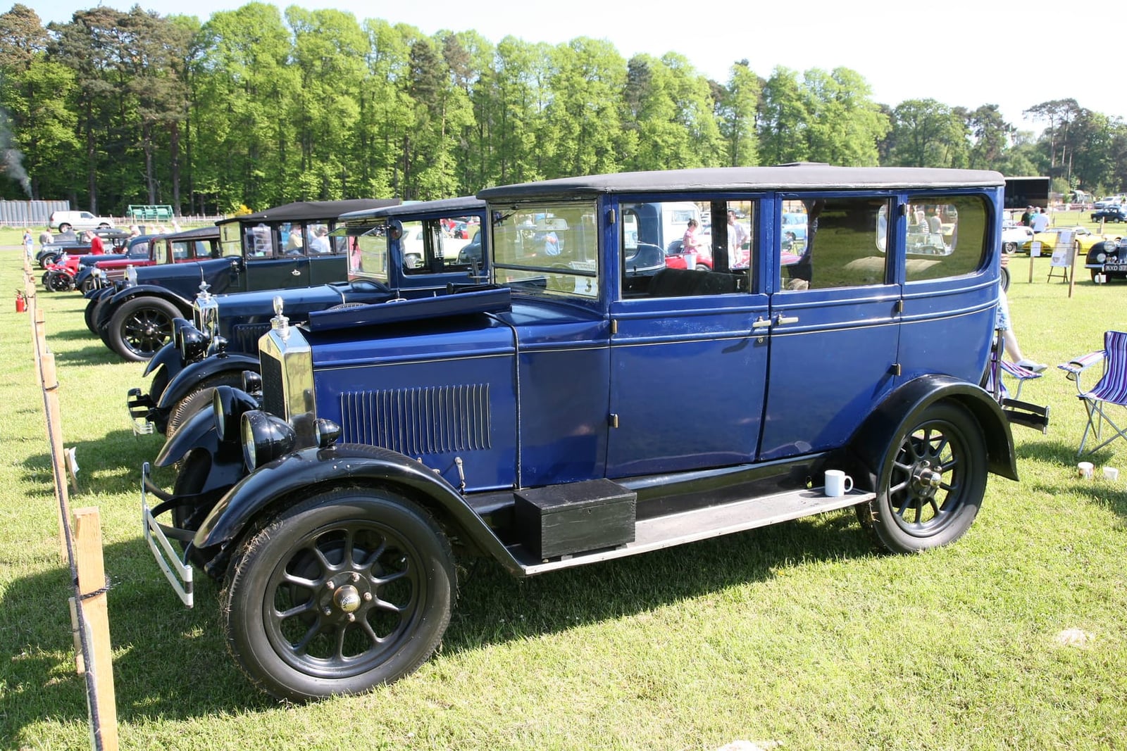 A dark blue 1927 Morris Oxford saloon with black wings on a showfield, other vintage cars behind